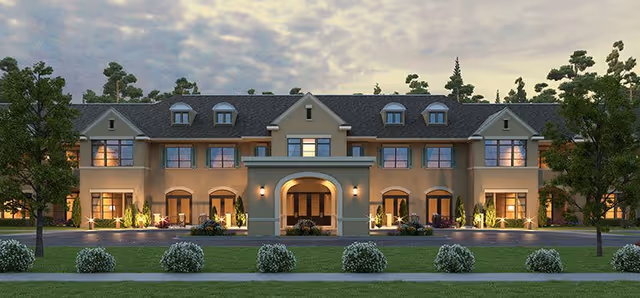 Front exterior view of a large, two-story senior living facility building with multiple windows and dormer windows on the roof. The building is illuminated with warm lighting and surrounded by landscaped greenery, including trees and bushes, under a cloudy sky at dusk.