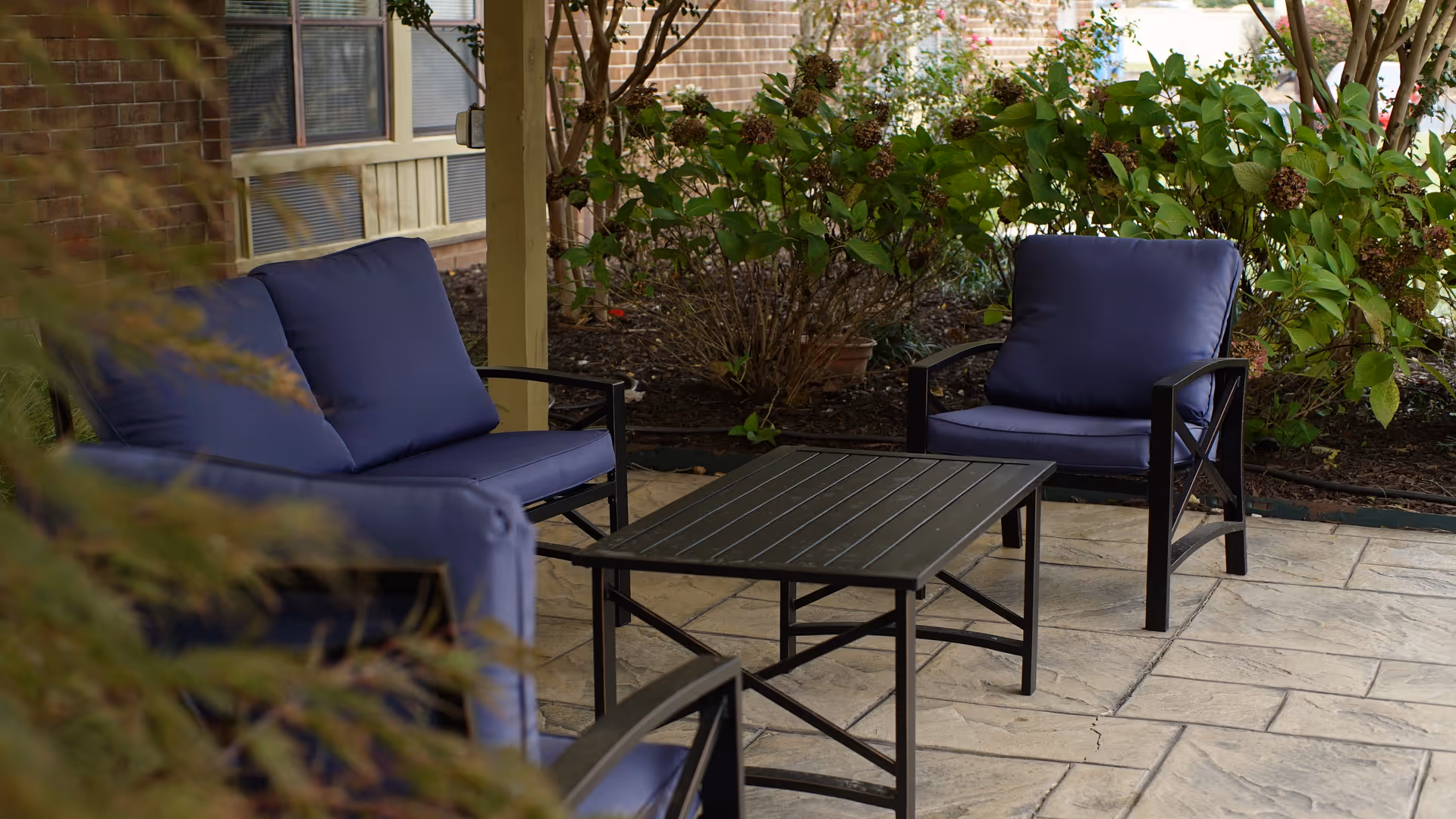 Outdoor patio area with a black metal table and matching chairs and loveseat with blue cushions, surrounded by plants and a brick building wall in the background.