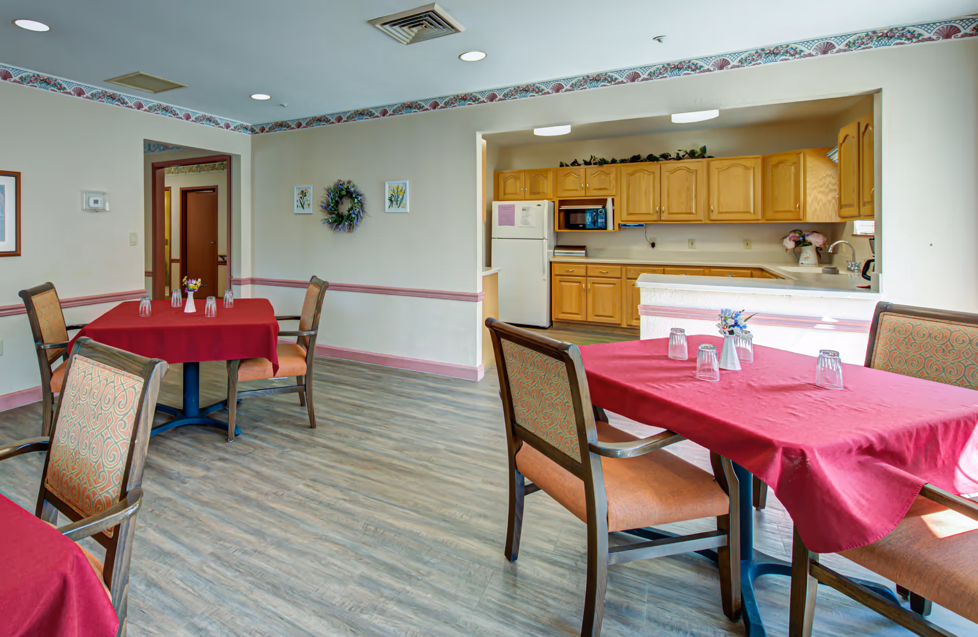 Dining area with tables covered in red tablecloths and chairs with patterned upholstery. The room has light-colored walls with decorative border trim and a wooden floor. In the background, there is a kitchen area with wooden cabinets, a refrigerator, microwave, and a sink. Small flower arrangements and upside-down glasses are placed on the tables.