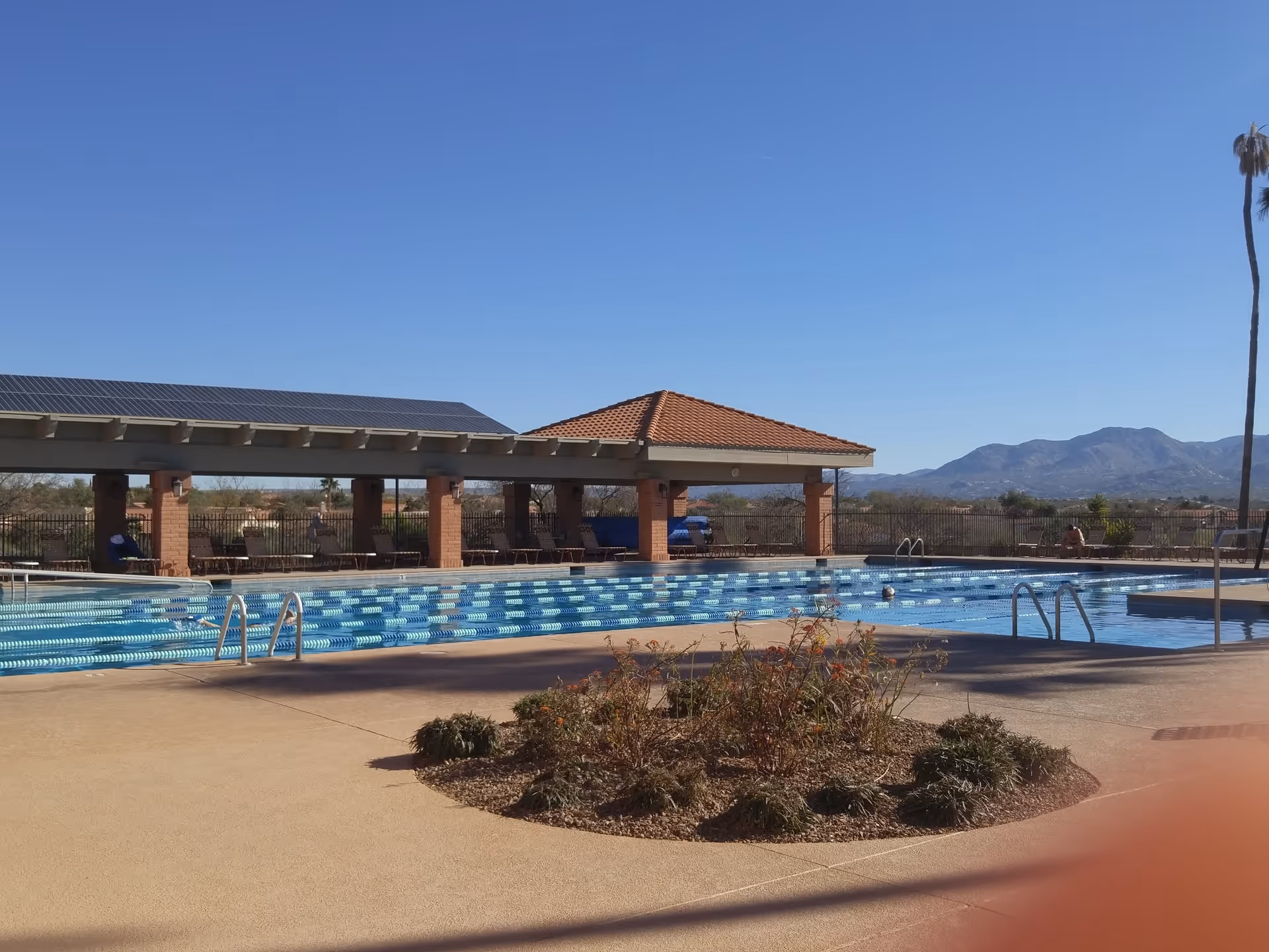 Outdoor swimming pool with lane markers and a covered pavilion, lounge chairs, and mountains in the background under a clear sky.