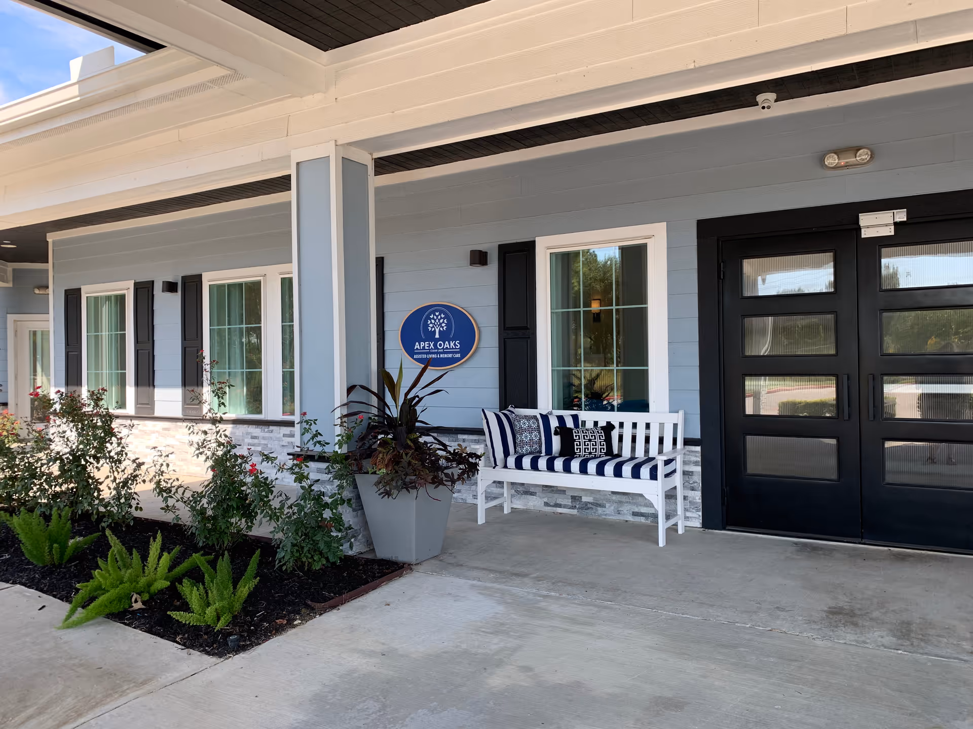 Entrance area of Apex Oaks at Clear Lake facility showing a covered porch with a white bench that has blue and white striped cushions and decorative pillows. There are several windows with black shutters, a large planter with plants, and a sign on the wall that reads 'Apex Oaks Assisted Living & Memory Care'. The exterior wall is painted light blue with stone accents near the bottom.