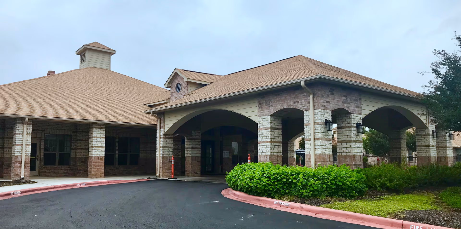 Exterior view of Mesa Vista Health Center showing a single-story building with a covered entrance supported by brick columns. The building has a tan roof and brick walls with some greenery and bushes near the entrance. The driveway curves around the entrance with red curbs marked with no parking and fire lane warnings.