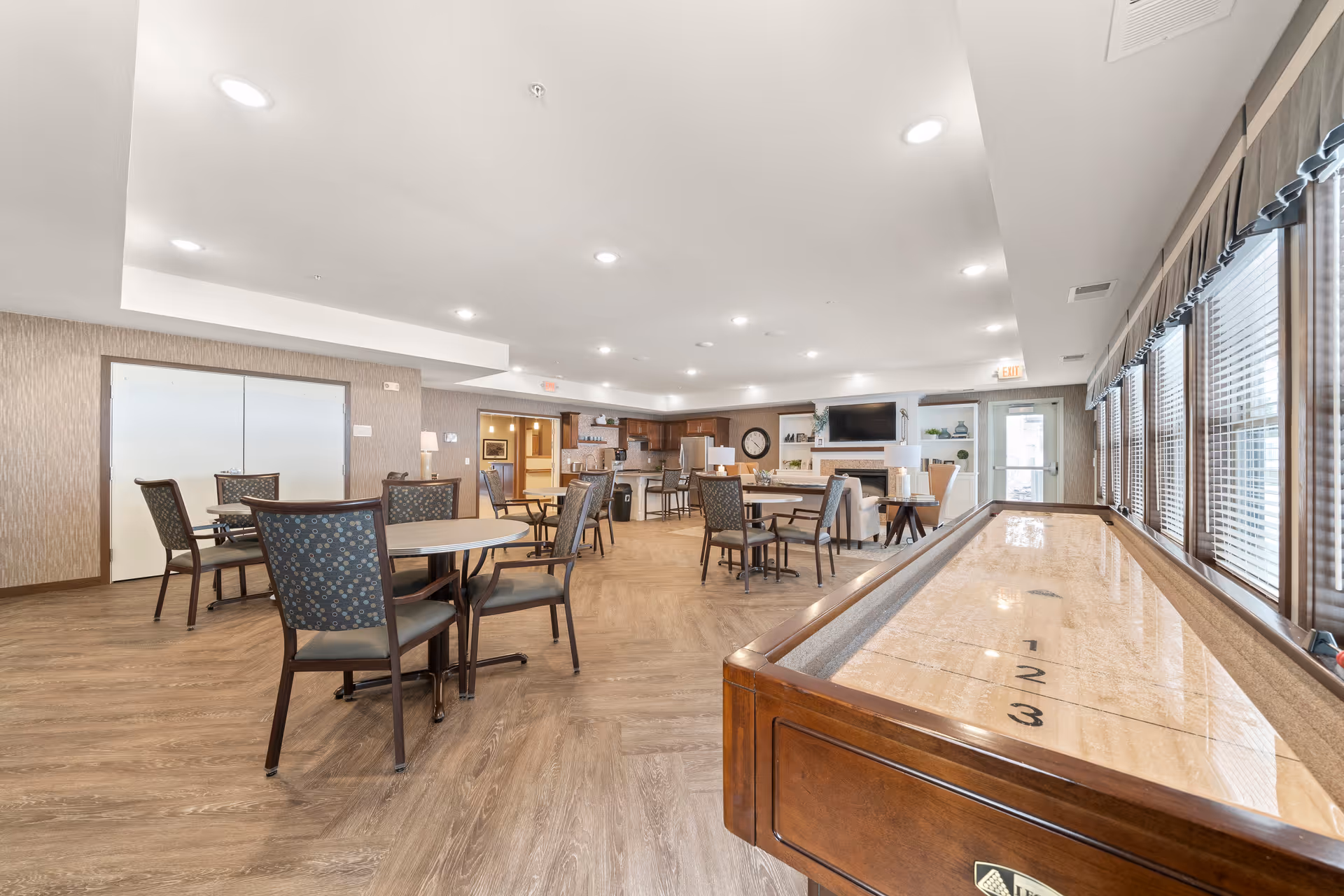Spacious senior living common area with tables and chairs, a shuffleboard table, and a TV-facing seating area near large windows.