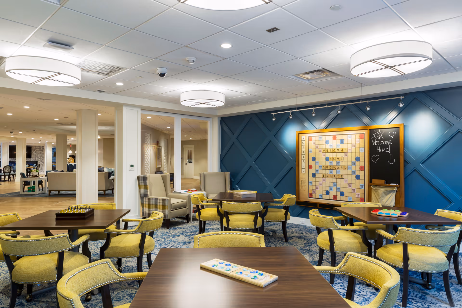 A well-lit common area with several tables and yellow cushioned chairs arranged for games. On the blue accent wall, there is a large Scrabble board and a chalkboard with the message 'Welcome Home!' along with drawings of a sun, hearts, and a smiley face. Comfortable armchairs are placed near the glass partition that separates this room from another seating area.
