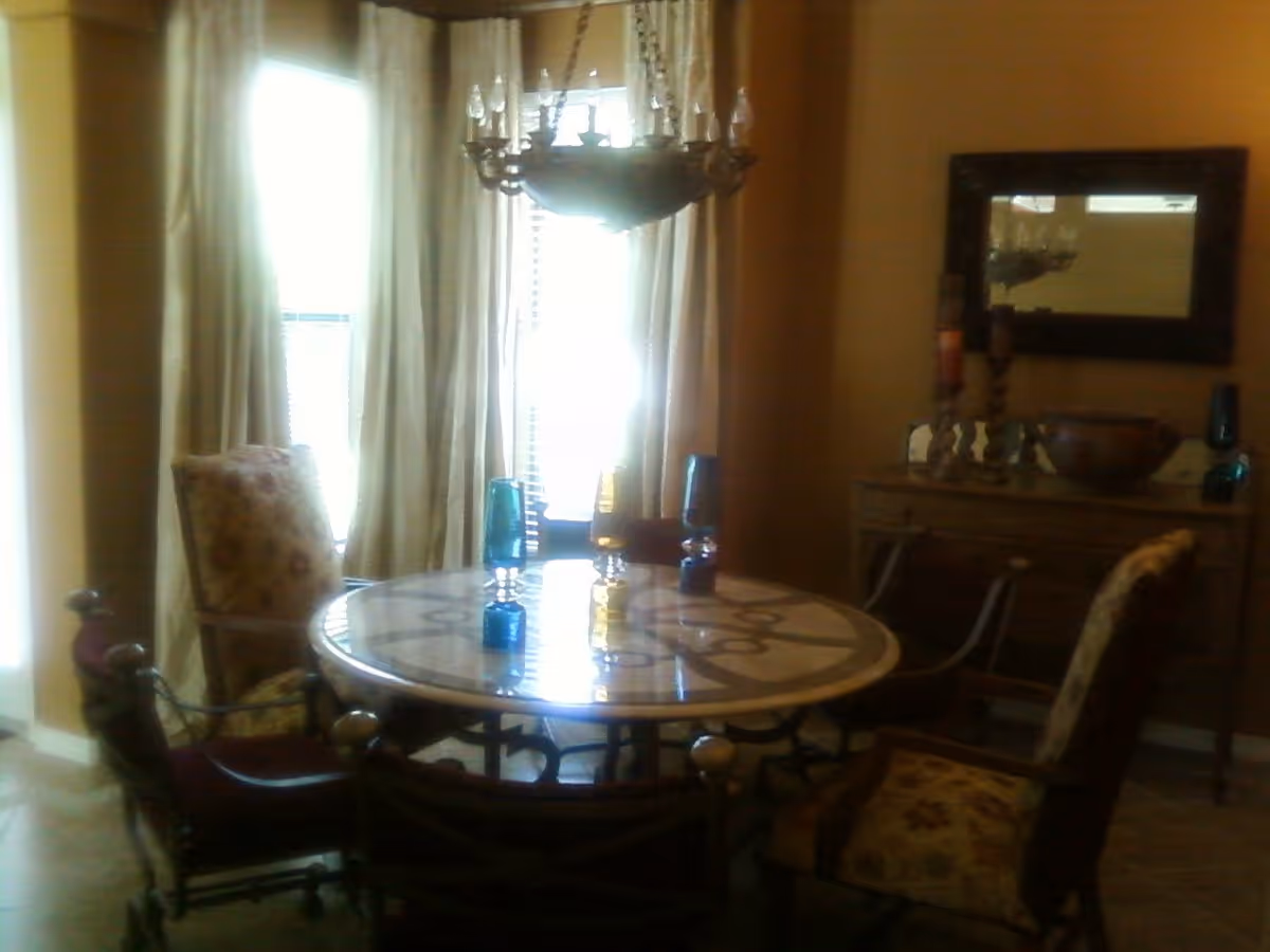 Round dining table with patterned chairs under a chandelier, tall curtained windows, and a sideboard with a mirror.