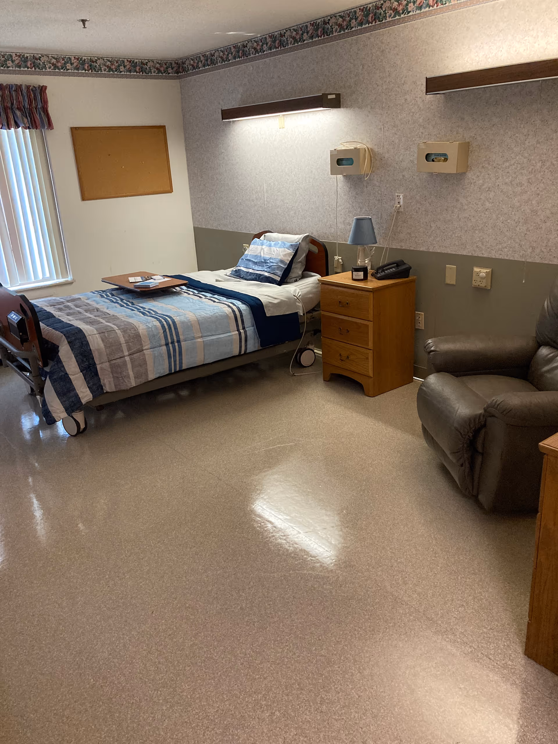 A senior living facility bedroom with a single hospital-style bed made with blue and gray striped bedding. Next to the bed is a wooden nightstand with a blue lamp, a phone, and a small candle. A brown recliner chair is positioned nearby. The room has a window with vertical blinds and a colorful valance, a cork bulletin board on the wall, and two wall-mounted tissue dispensers above the nightstand.