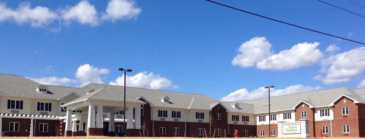 Exterior view of a large two-story senior living facility building under a blue sky with scattered clouds. The building has a combination of red brick and white siding with multiple windows and a covered entrance supported by white columns. There are street lamps and a sign in front that reads 'English Meadows'.
