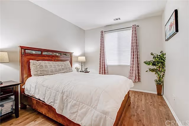 A bedroom with a wooden bed frame and white bedding, a large pillow, two bedside tables with lamps, a window with patterned curtains, a potted plant in the corner, and a framed picture on the wall.