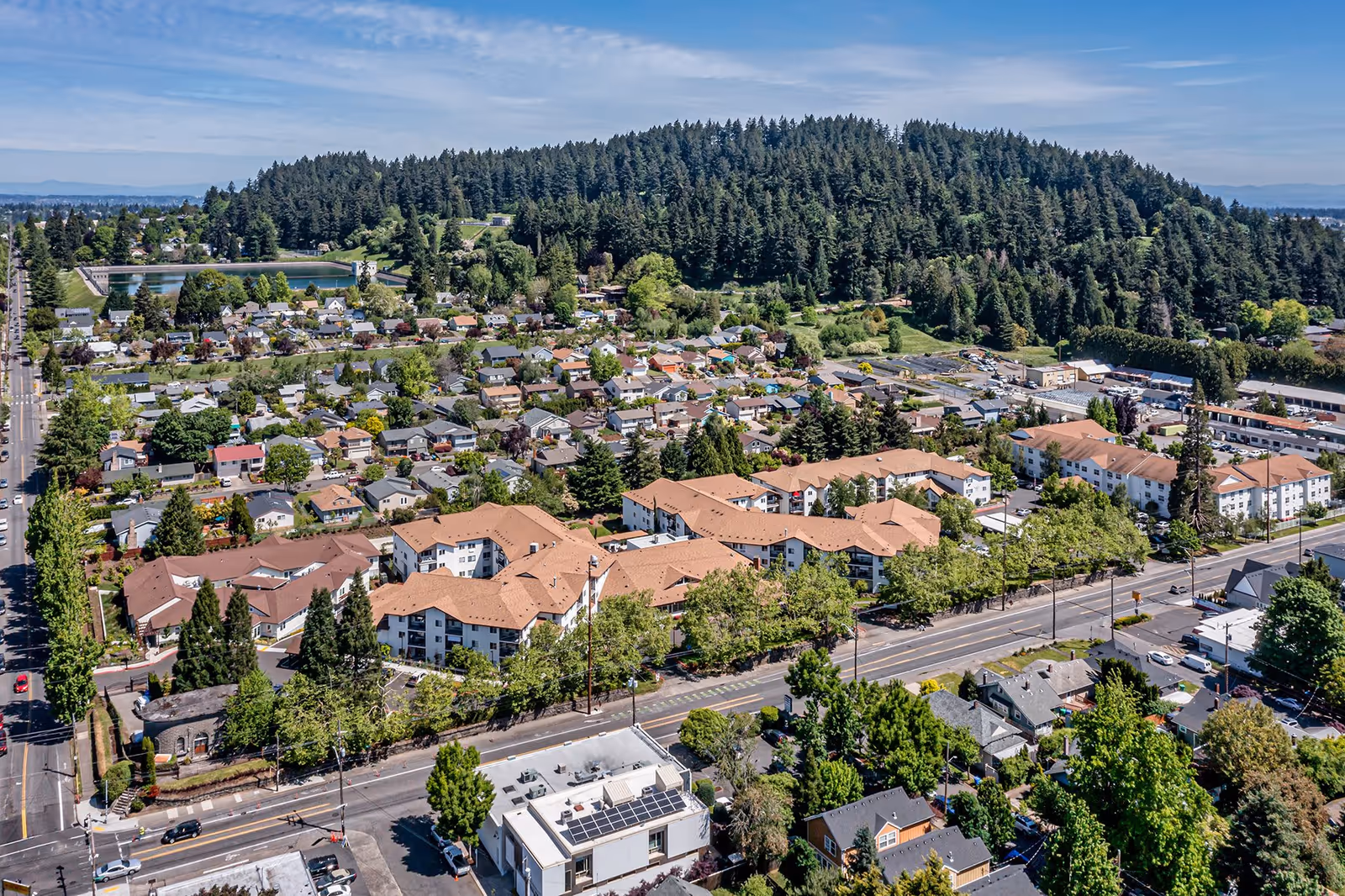 Aerial view of a residential neighborhood with a large senior living facility featuring multiple connected buildings with tan roofs in the center. Surrounding the facility are houses, trees, and roads, with a forested hill in the background under a partly cloudy sky.