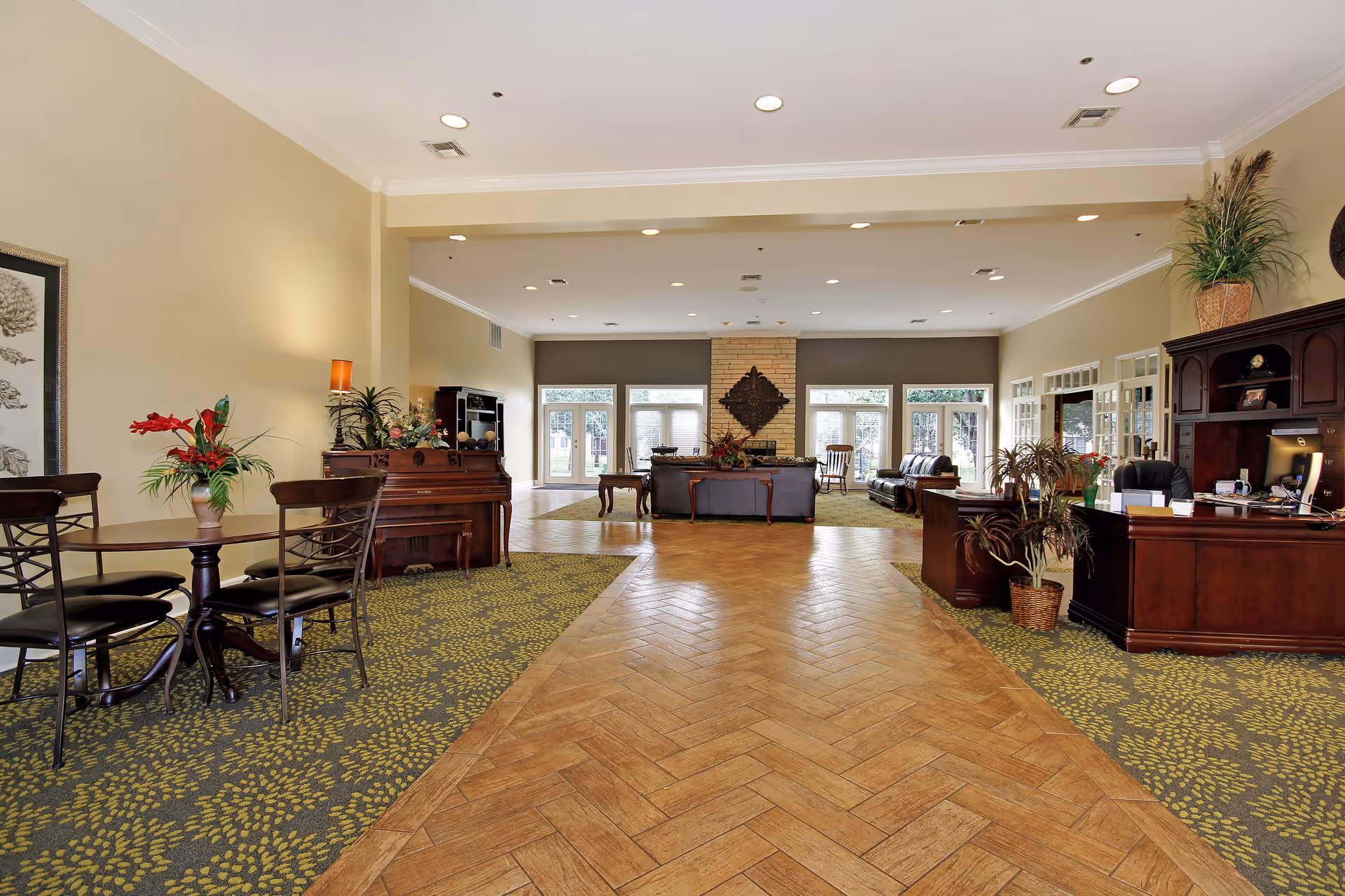 Spacious senior living facility common area with a wooden floor pathway leading to a seating area with leather sofas and a fireplace. The room features large windows with views of greenery outside, a piano with floral arrangements, a small table with chairs, and a wooden reception desk with plants and office equipment.