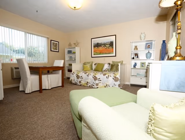 A cozy living room area with a floral-patterned daybed adorned with green and beige pillows, a green armchair with a matching ottoman in the foreground, a wooden table with two white slipcovered chairs near a window with vertical blinds, and white shelving units displaying decorative items and framed photos. The walls are beige and there is a framed landscape painting above the daybed.