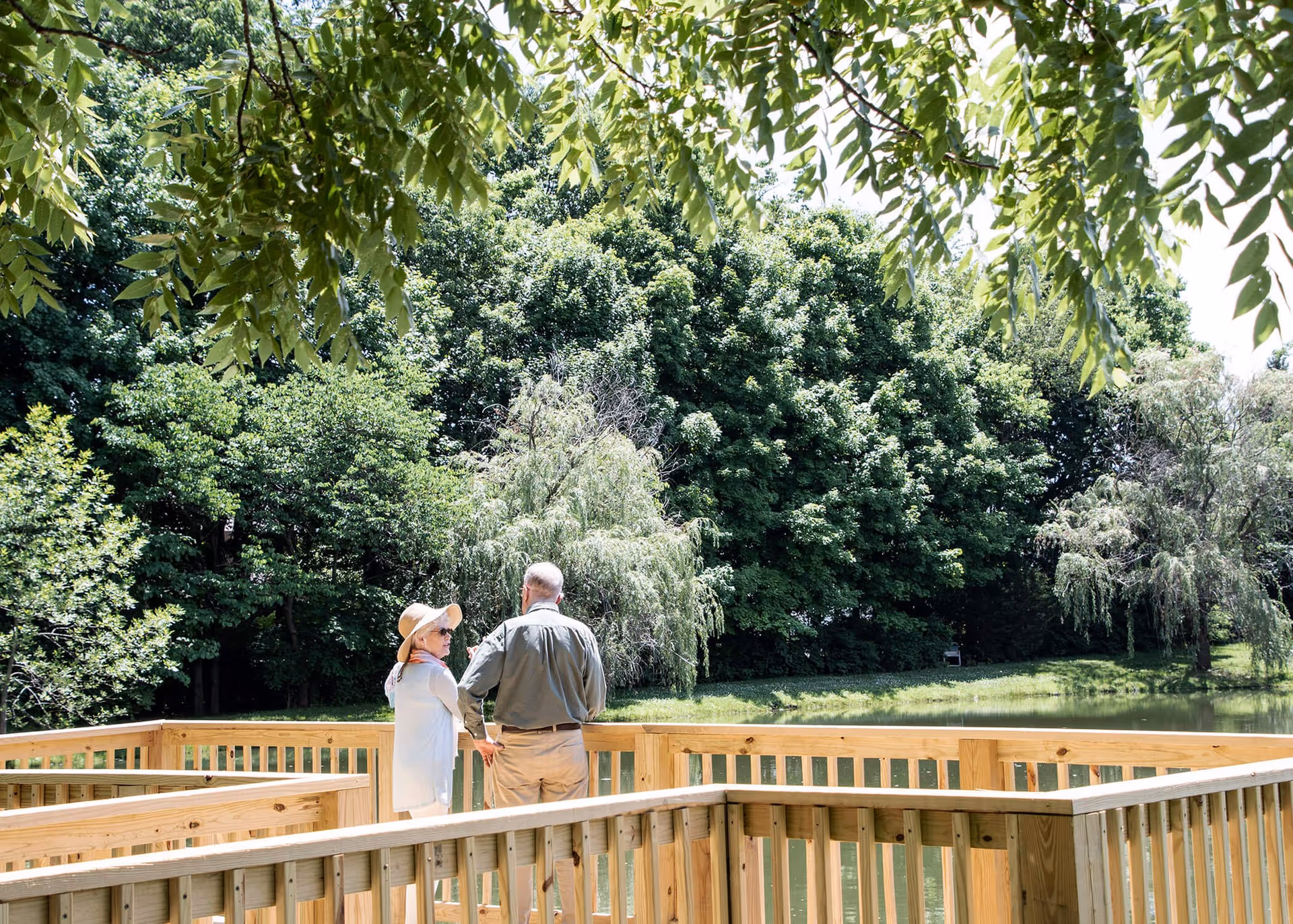 An elderly couple stands on a wooden deck overlooking a pond surrounded by lush green trees on a sunny day. The woman wears a sunhat and light clothing, while the man is dressed in a long-sleeve shirt and pants. Overhanging tree branches frame the top of the image.