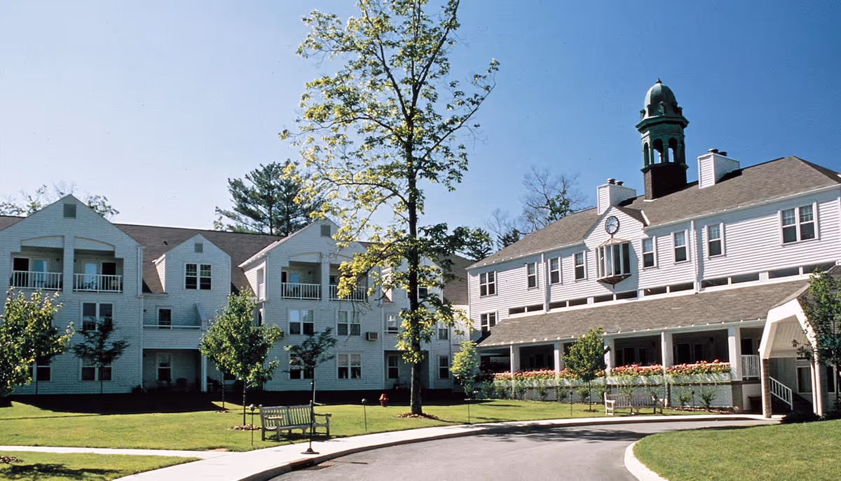 Exterior view of The Fountains at Millbrook senior living facility showing white multi-story buildings with balconies, a clock tower, green lawn, trees, benches, and a clear blue sky.