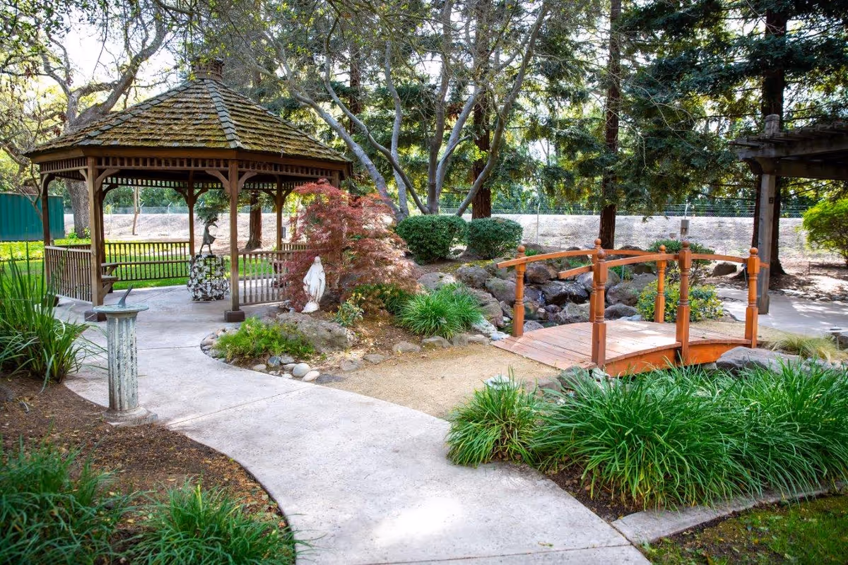 A peaceful outdoor garden area featuring a wooden gazebo with a shingled roof, a small wooden bridge over a rocky stream, various green plants and bushes, a statue, and a concrete pathway winding through the scene.