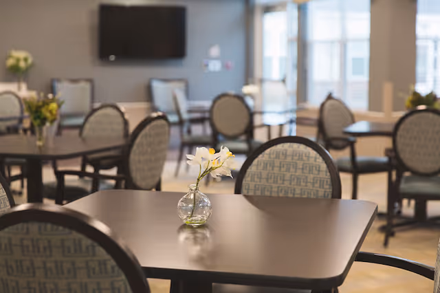 Bright assisted living dining room with dark wooden tables, patterned round-backed chairs, and small vases of flowers on the tabletops.