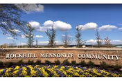 Stone sign reading 'Rockhill Mennonite Community' with a flower bed of yellow and purple flowers in front, trees and a blue sky with scattered clouds in the background.