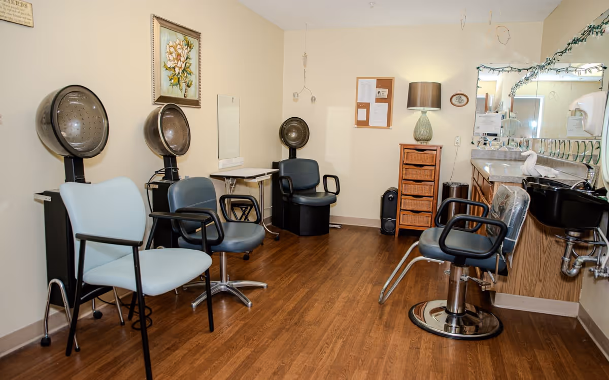 Interior view of a salon area in a senior living facility with multiple salon chairs, hair dryers, a wooden floor, a lamp on a wicker drawer unit, a large mirror above a countertop with a sink, and a framed floral painting on the wall.
