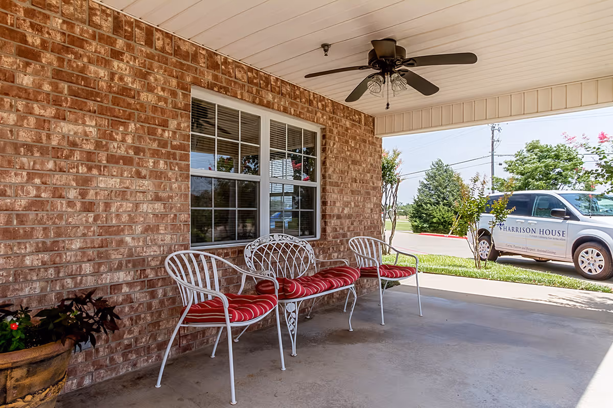 Covered outdoor patio area with a brick wall, a window, and three white metal chairs with red cushions. A ceiling fan is mounted on the white ceiling. Outside, there is a parking area with a van labeled 'Harrison House' and some trees and greenery in the background.