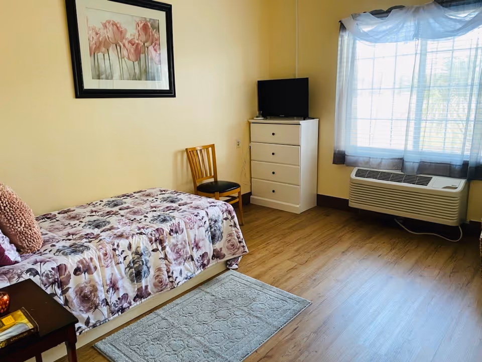 Small bedroom with a single bed covered in a floral bedspread, a dresser with a TV, a wooden chair, a window with curtains and an air conditioning unit.