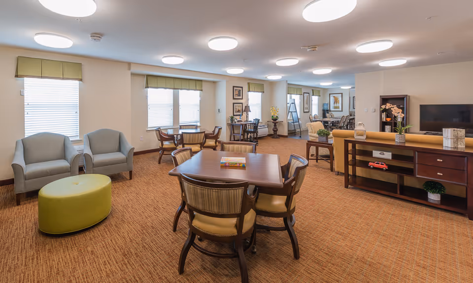 A spacious and well-lit common area in a senior living facility featuring multiple seating arrangements including a table with four chairs, two armchairs with a green ottoman, and a sofa facing a television. The room has beige walls, carpeted floor, several windows with green valances, and ceiling lights. Decorative plants and framed pictures adorn the space.