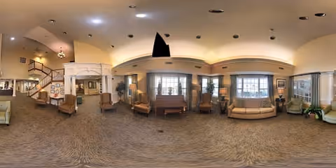 A spacious and well-lit living room area with multiple seating options including armchairs and sofas arranged around a central piano. Large windows allow natural light to fill the room, and there are plants and framed pictures decorating the space. A staircase is visible on the left side of the room.