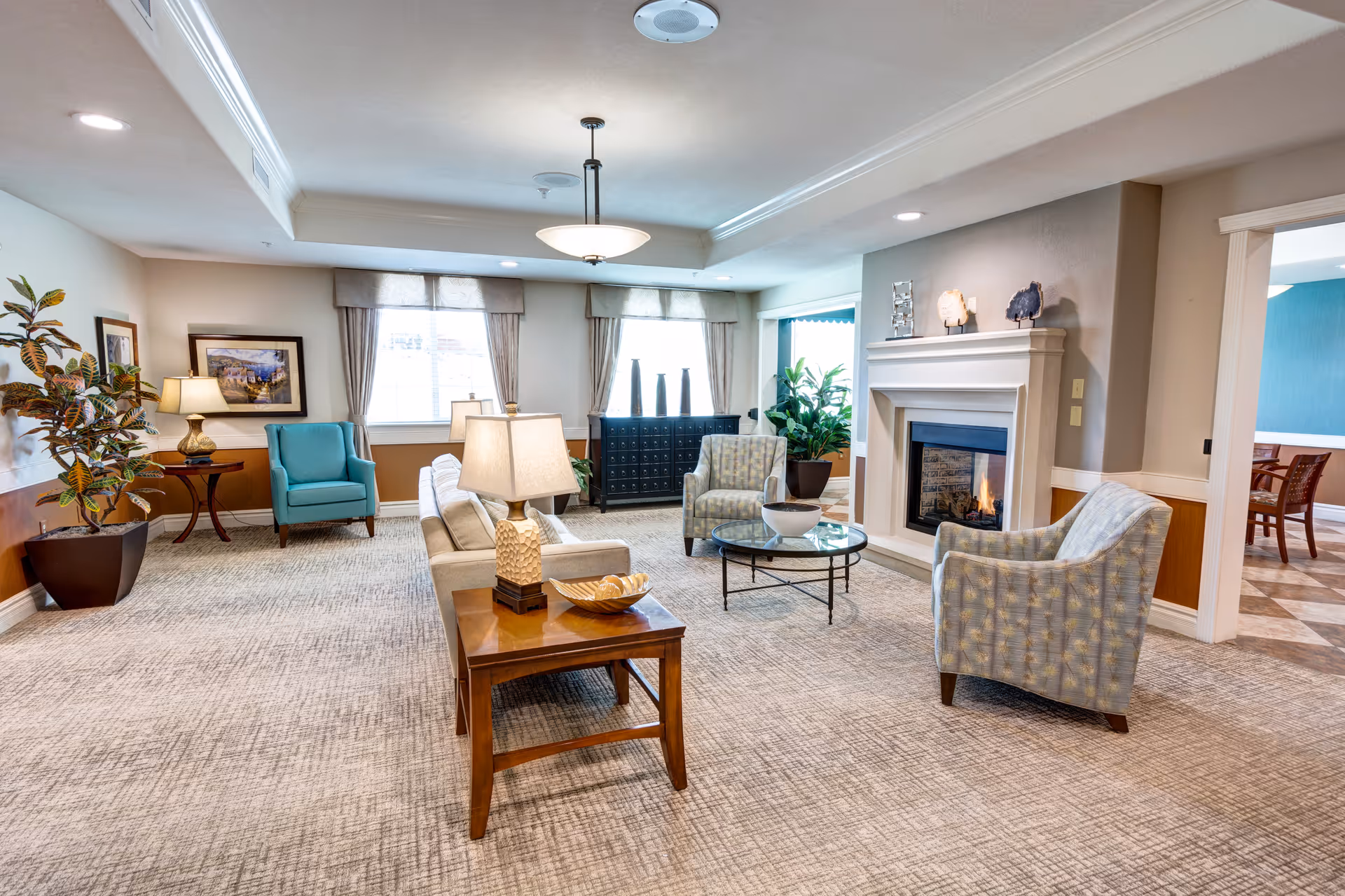 A bright and spacious senior living facility common area with comfortable seating including armchairs and a sofa, a glass coffee table, a fireplace, decorative plants, framed artwork on the walls, and large windows with curtains allowing natural light to fill the room.