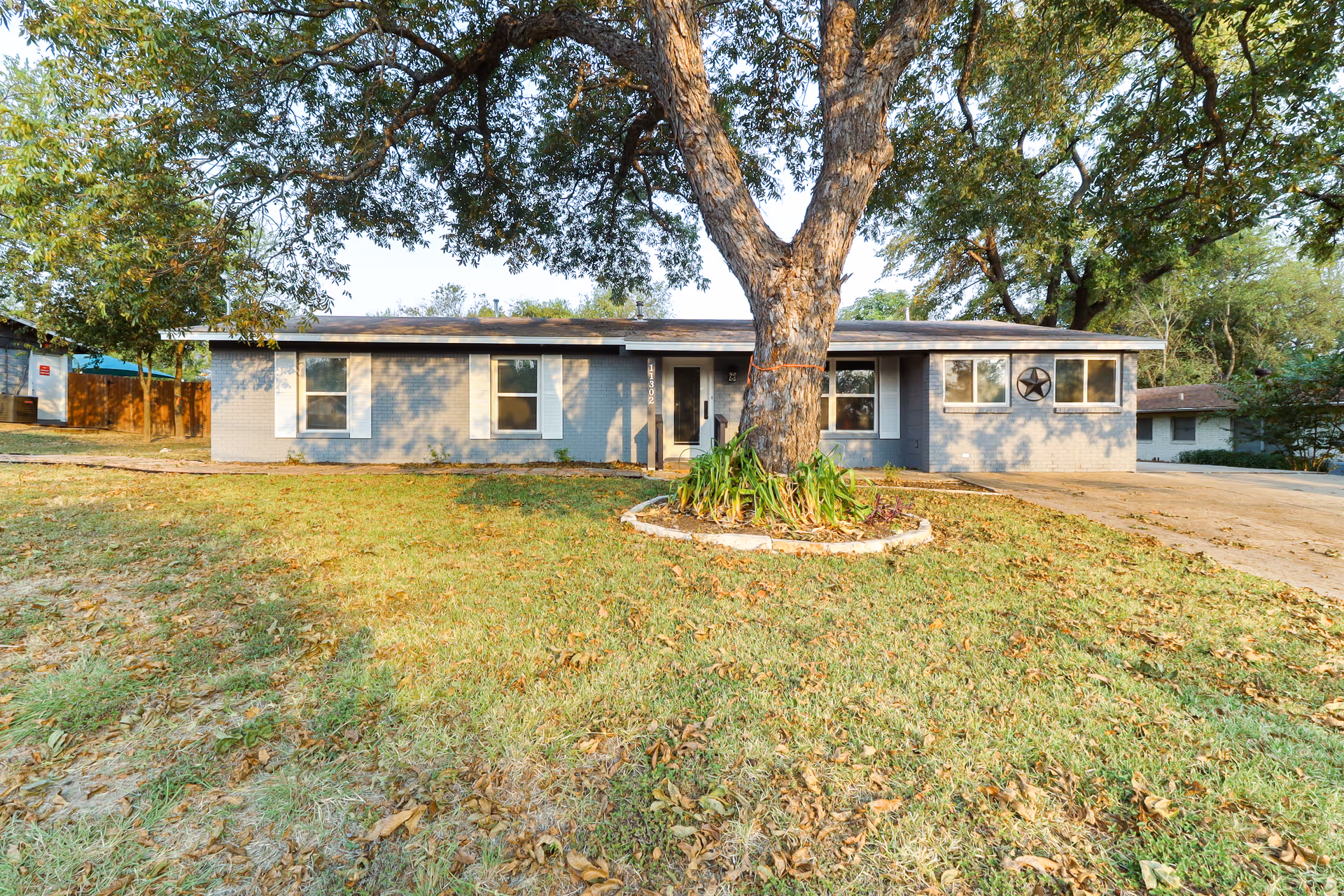 Single-story gray brick building with white window shutters and a large tree in the front yard, surrounded by grass and some fallen leaves. The building has a driveway on the right side and a star decoration on the wall near the windows.