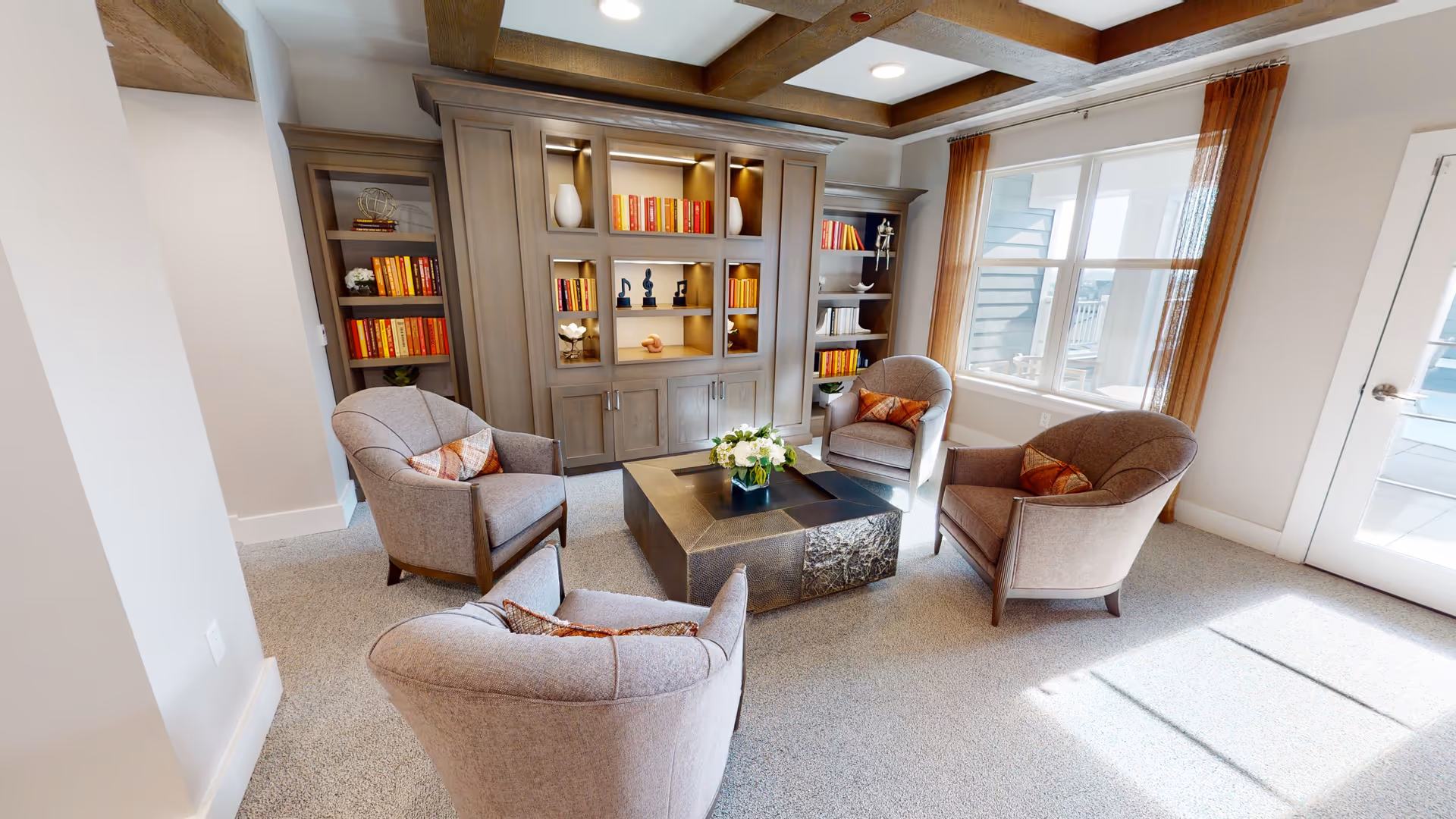 Bright communal living room with four armchairs arranged around a central coffee table, built-in bookcases, and large windows.
