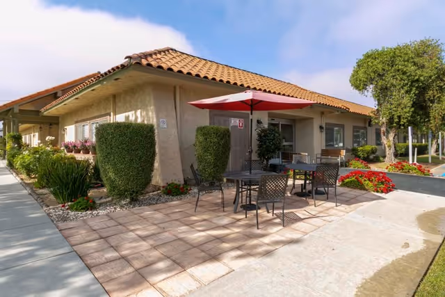 Outdoor patio area of a healthcare facility with a tiled roof building, patio tables and chairs, a red umbrella, neatly trimmed bushes, and colorful flower beds under a partly cloudy sky.