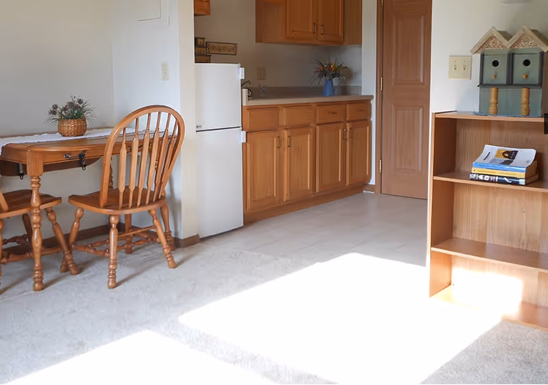 A small kitchen area with wooden cabinets, a white refrigerator, and a wooden table with a matching chair. There is a small potted plant on the table and a bookshelf with books and decorative birdhouses on top. Sunlight is streaming onto the carpeted floor.