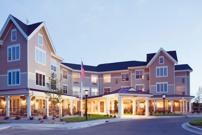 Exterior view of a large, multi-story assisted living facility building with beige siding and white trim, featuring multiple windows and a covered entrance with benches. The American flag is displayed on a flagpole near the entrance, and the scene is set during early evening with lights glowing warmly from inside the building.