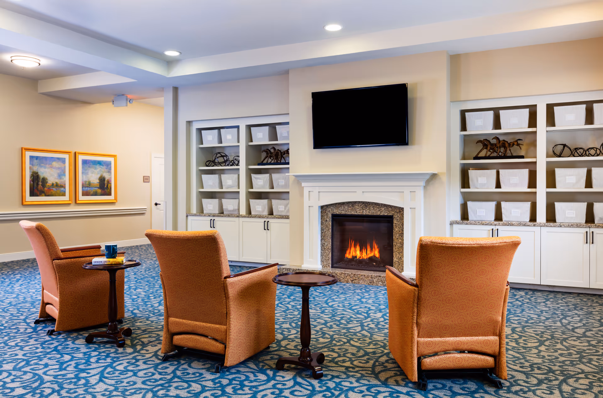 A cozy common area with three orange upholstered armchairs arranged around two small wooden tables, a lit fireplace with a granite surround, a wall-mounted flat screen TV above the fireplace, built-in white shelves filled with white storage baskets and decorative items, and two framed landscape paintings on the beige wall.