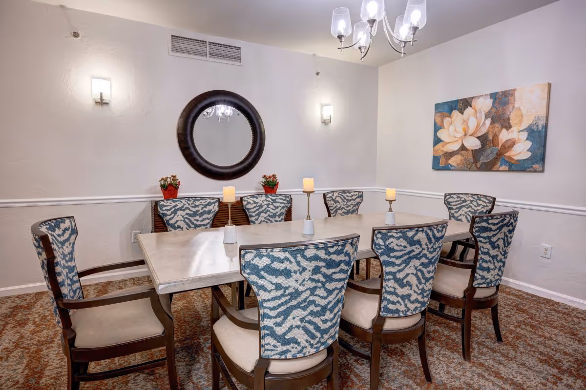 A dining room with a rectangular table surrounded by eight upholstered chairs with blue and white patterned backs. On the table are four candle holders with lit candles. The room has beige walls with white trim, a round mirror on one wall, two small potted plants, a floral painting on another wall, and a chandelier hanging from the ceiling.
