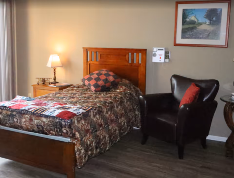 A furnished bedroom with a single wooden bed, patterned quilt, bedside table and lamp, and a leather armchair under wall art.