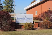 Sign reading 'Sunrise Terrace Nursing and Rehabilitation Center' on a landscaped lawn in front of a red brick building.