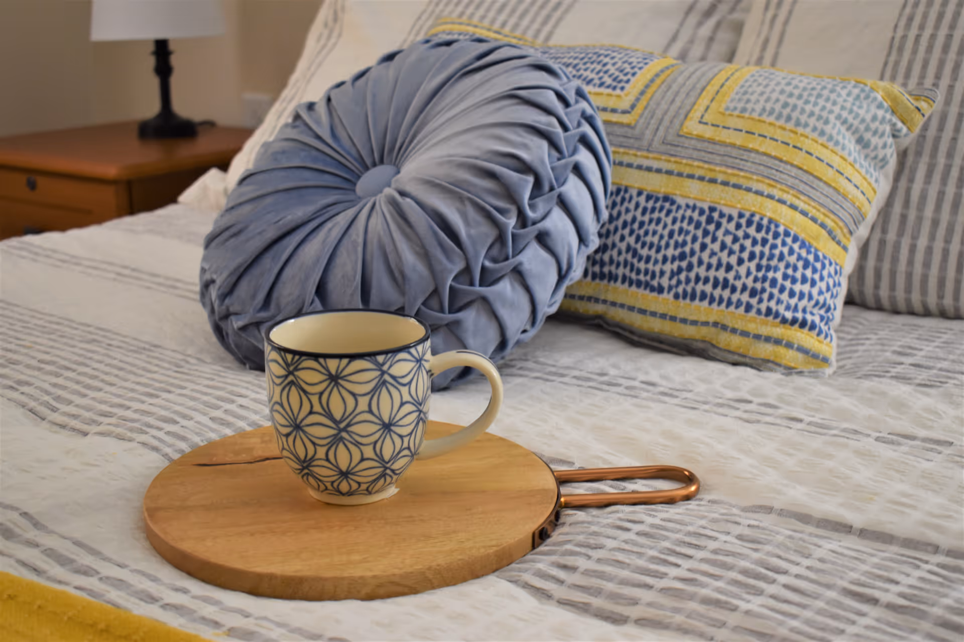 A patterned mug on a wooden serving board sits on a neatly made bed with decorative pillows and a bedside lamp in the background.