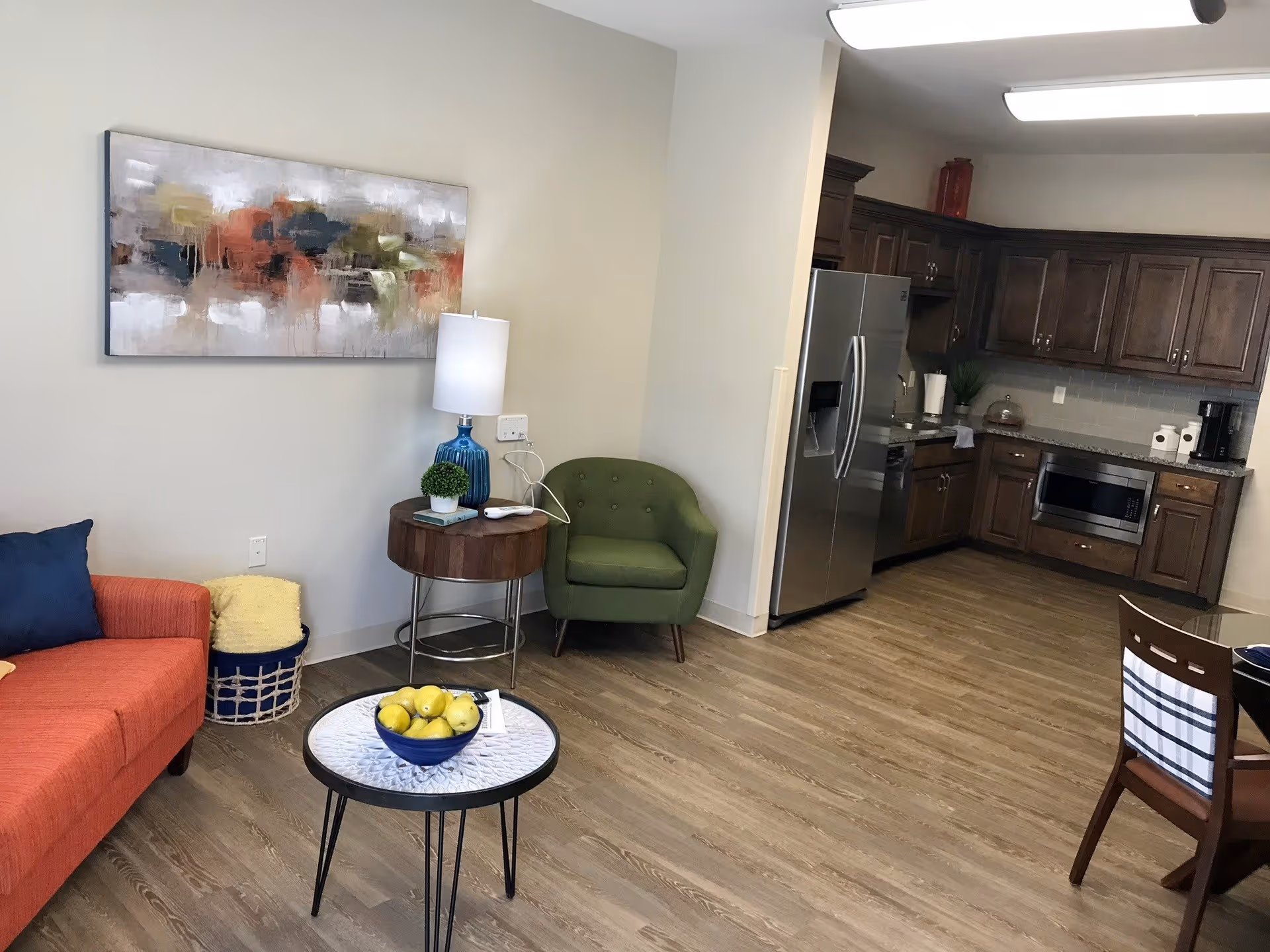 A cozy living area with an orange sofa, a small round table with a bowl of yellow apples, a green armchair, and a side table with a blue lamp and a small plant. In the background, there is a kitchen with wooden cabinets, a stainless steel refrigerator, microwave, and coffee maker. The floor is wooden and there is a colorful abstract painting on the wall.