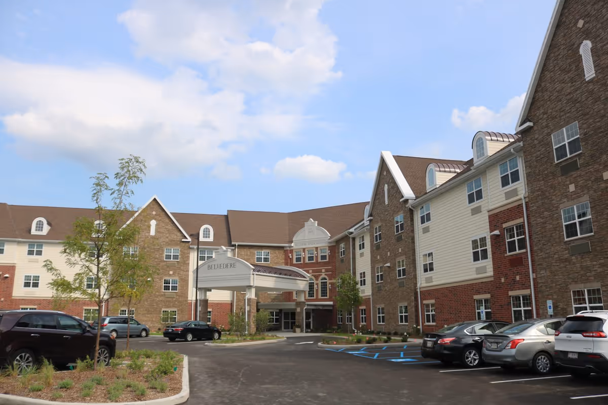 Exterior view of Belvedere Senior Housing building with a large covered entrance, multiple windows, and a parking lot with several cars parked. The building has a mix of brick and siding with a brown roof under a partly cloudy sky.