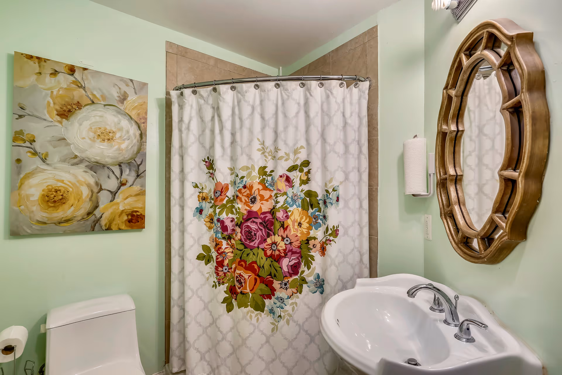 Bathroom with a white toilet, a white sink with silver faucets, a decorative wooden-framed mirror, a floral shower curtain with colorful flowers, and a painting of white and yellow flowers on a light green wall.
