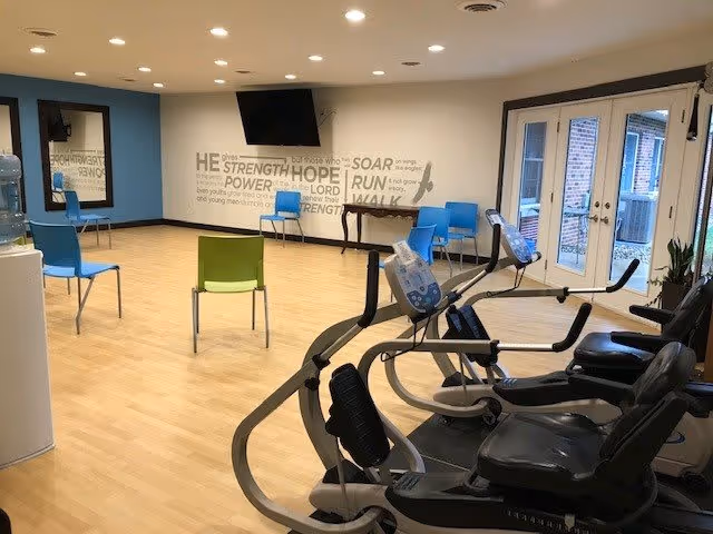 Bright interior exercise/activity room with recumbent bikes, scattered chairs, a wall-mounted TV, and glass French doors.