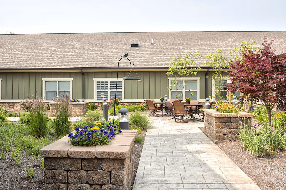Outdoor patio area at Dominion Senior Living of Anderson featuring a paved walkway, raised stone flower beds with colorful flowers, several small trees and shrubs, and a seating area with tables and chairs against the backdrop of a green building with white-trimmed windows.
