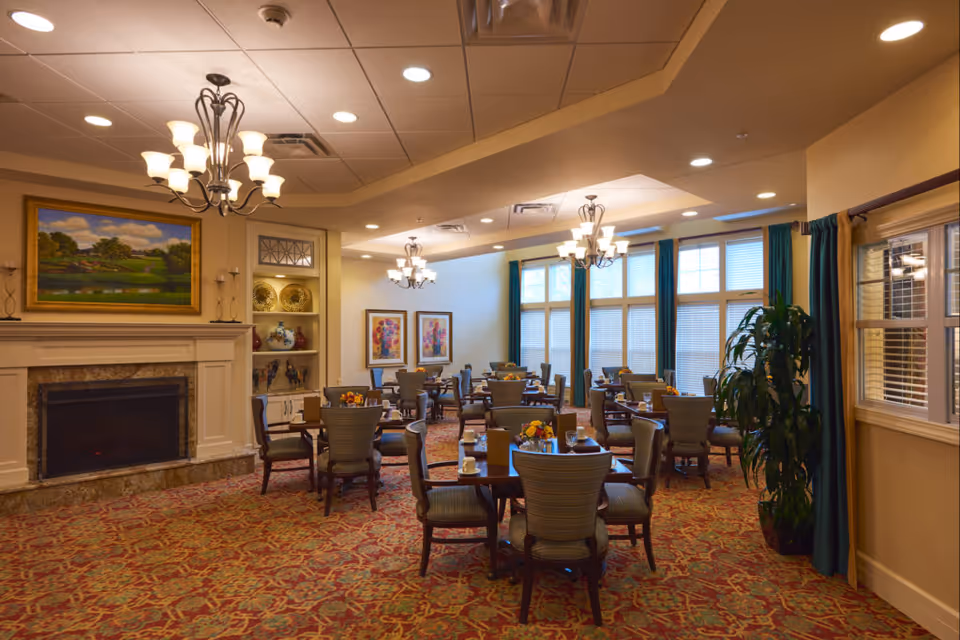 A warm and inviting dining room with multiple round tables and chairs arranged neatly. The room features a fireplace with a landscape painting above it, decorative shelves with vases and plates, large windows with teal curtains, and elegant chandeliers hanging from the ceiling. The carpet has a red and gold pattern, and the room is well-lit with recessed lighting.