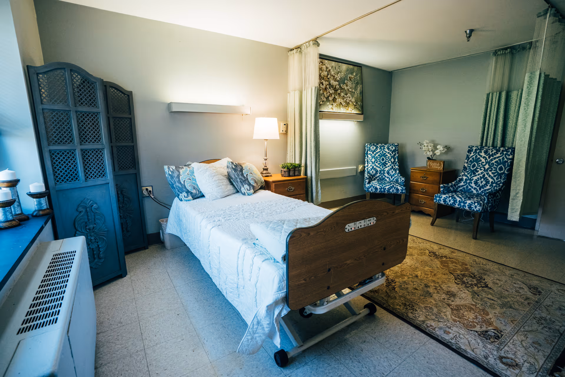 A well-lit bedroom in a senior living facility with a single hospital-style bed covered in white bedding and decorative pillows. Next to the bed is a wooden nightstand with a lamp and small potted plants. Two blue patterned chairs and a wooden chest of drawers are placed against the wall, with a floral painting hanging above. A decorative room divider is positioned near the window, and a patterned area rug covers part of the floor.
