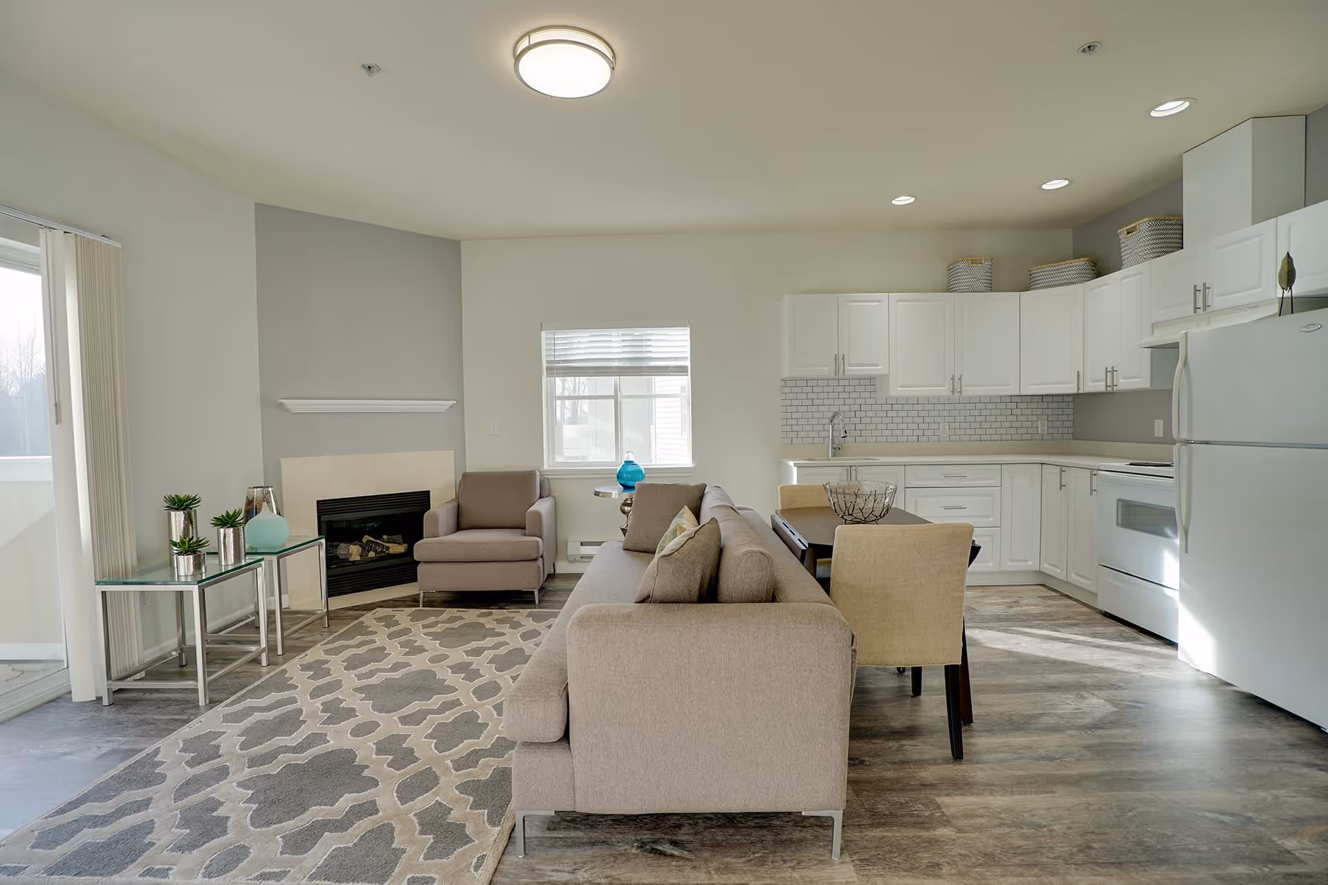 A bright and modern open-concept living room and kitchen area in a retirement facility. The living room features a beige sofa with cushions, a matching armchair, a patterned area rug, and a glass side table with decorative plants and a vase. The kitchen has white cabinets, a white refrigerator, stove, and a dining table with beige chairs. A window and sliding glass door allow natural light to fill the space.