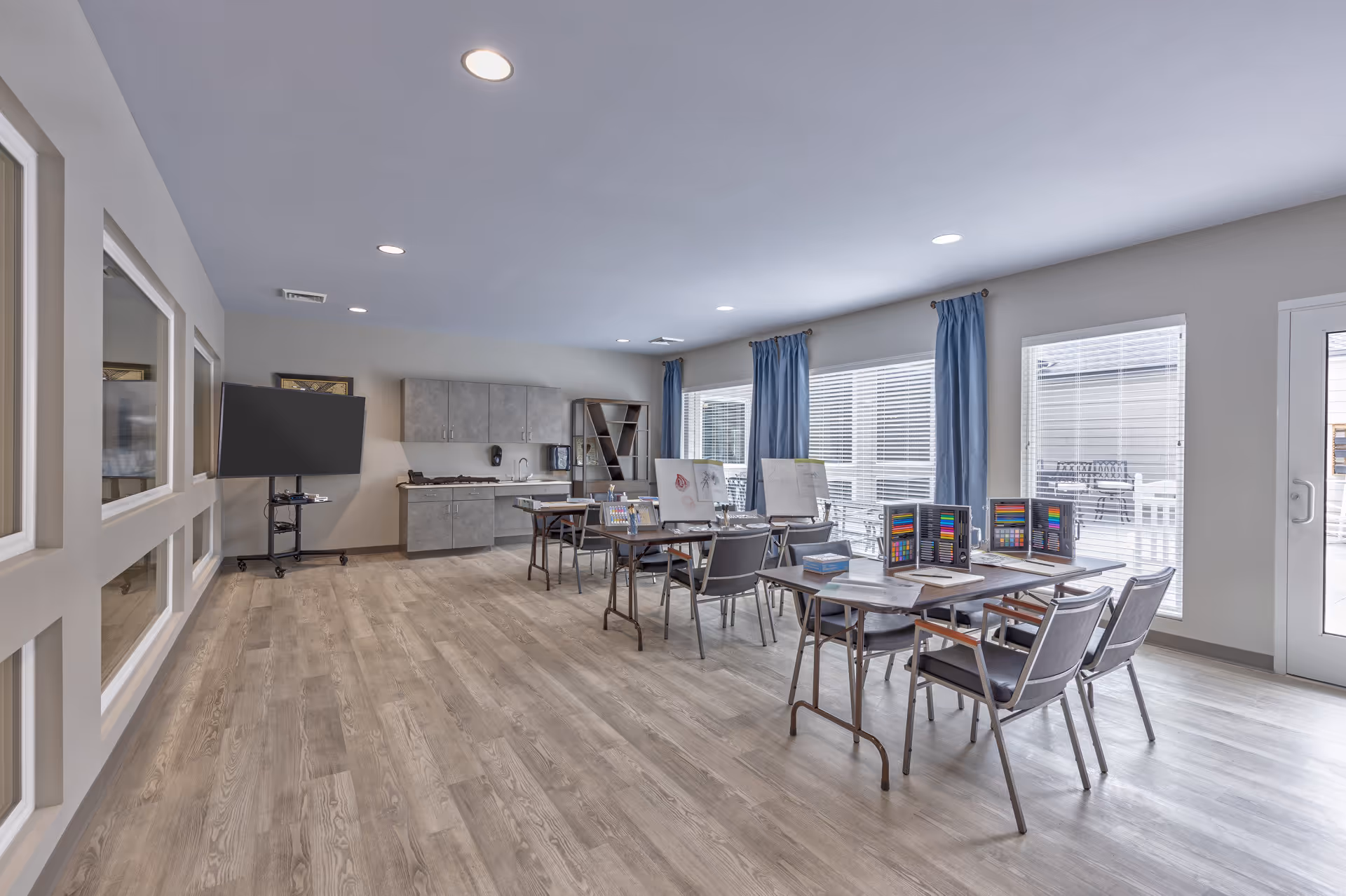 A bright, spacious activity room with wooden flooring and large windows covered with blue curtains. Several tables and chairs are arranged with art supplies, including colored pencils and drawing pads. A large flat-screen TV is mounted on a stand near the wall, and cabinets with a sink are visible in the background.