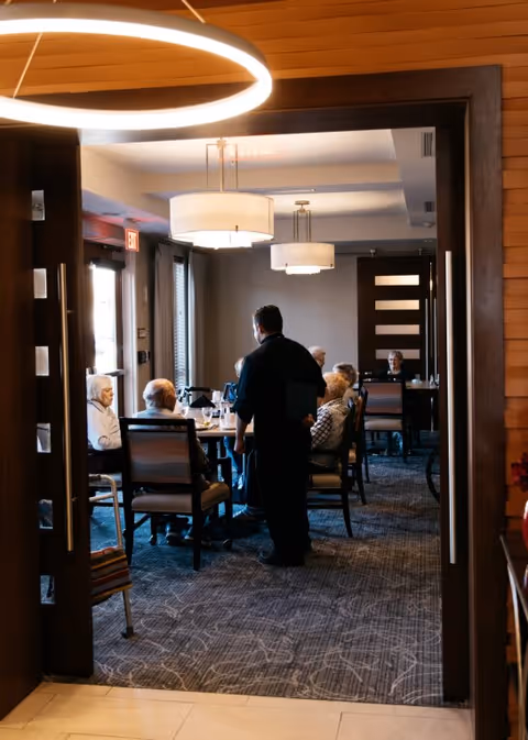 A group of elderly people seated around a dining table in a well-lit room, with a staff member standing and attending to them. The room has modern light fixtures and large windows allowing natural light.