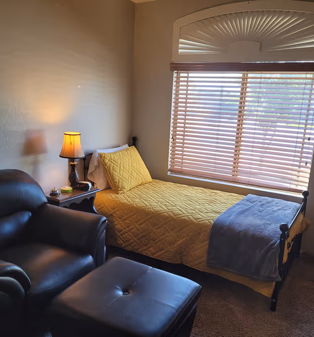 A cozy bedroom with a single bed covered in a yellow quilt and a gray blanket at the foot. Next to the bed is a small nightstand with a lamp, an alarm clock, and a small container. A large window with wooden blinds lets in natural light. In the foreground, there is a black leather armchair and matching ottoman.