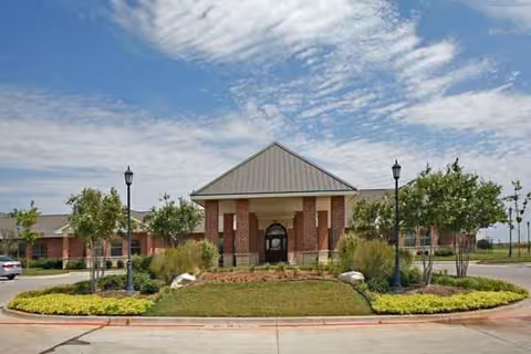 Front of a brick senior facility with landscaped entrance under a blue sky.