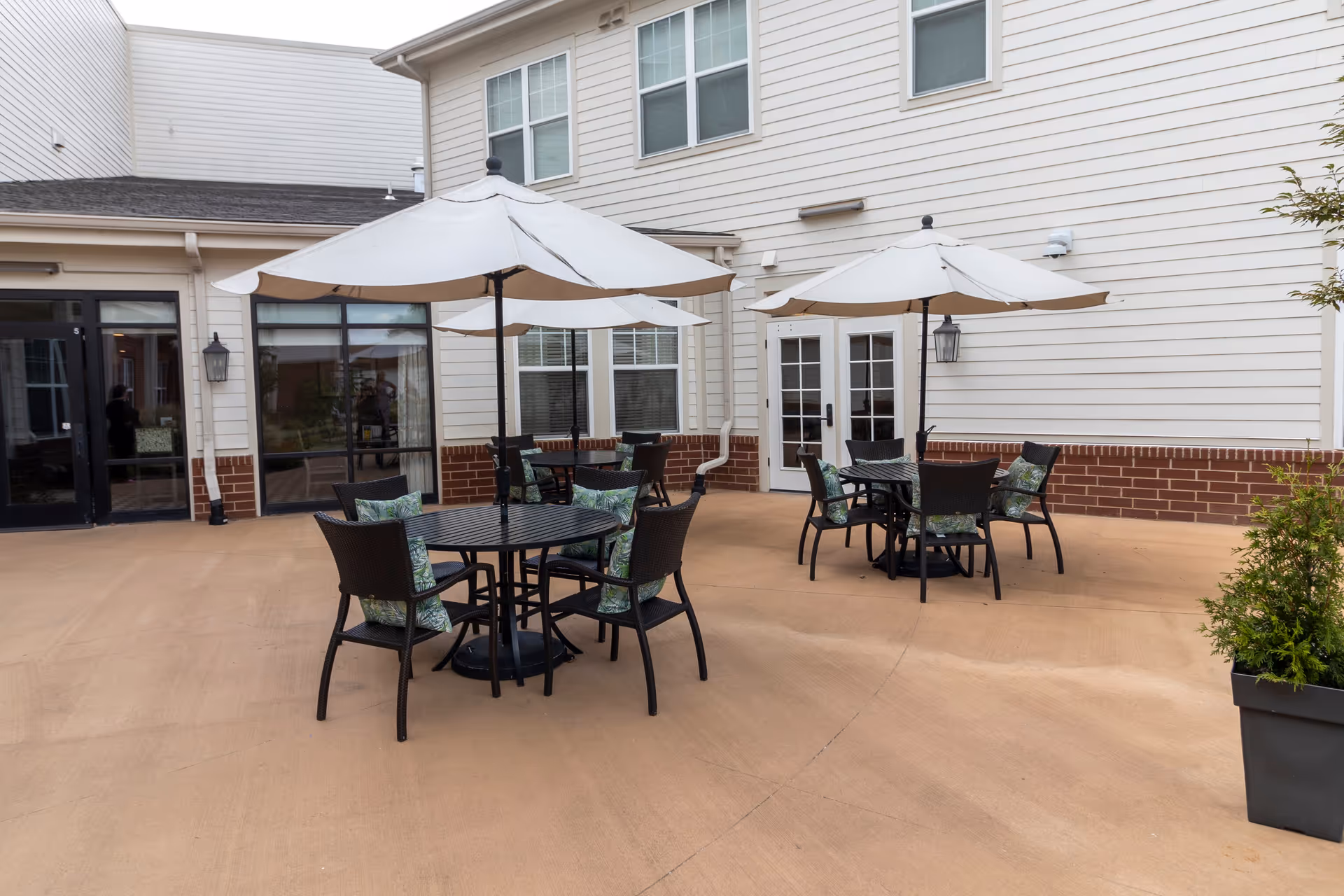Outdoor patio area with three round black tables, each surrounded by four black chairs with green patterned cushions. Each table has a large white umbrella providing shade. The patio is adjacent to a building with white siding and brick trim, featuring multiple windows and doors.