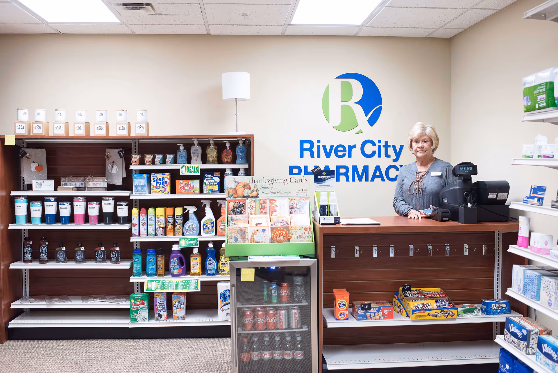 A woman stands behind the counter of a small pharmacy area labeled River City Pharmacy. Shelves behind and beside her display various household and personal care products, greeting cards, and beverages in a small refrigerator. The setting appears clean and well-organized with a neutral wall color and ceiling lights.